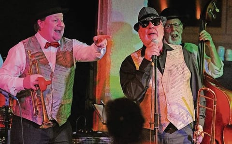 Musicians performing on stage under colored lighting, with performers in vests and hats singing into microphones