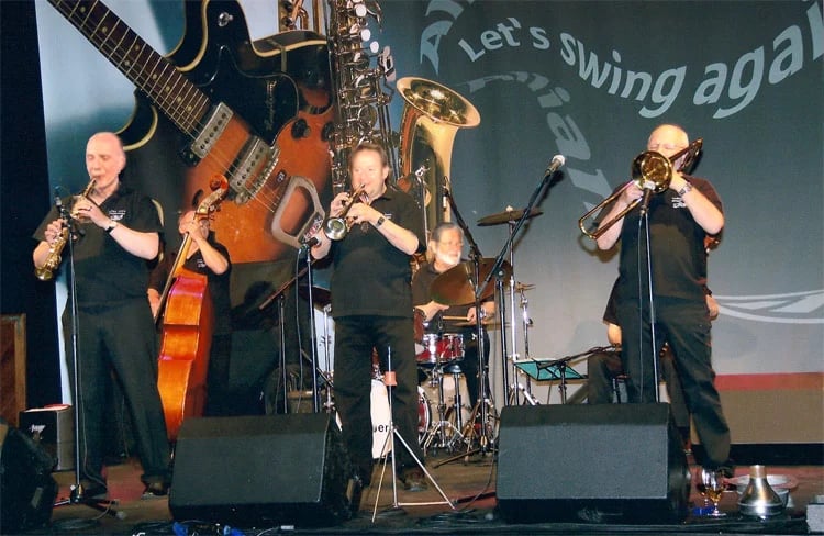 Jazz band performing on stage with musicians playing saxophone, trombone, guitar, and cello instruments.
