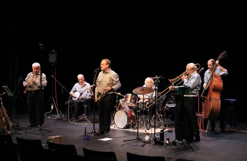 A jazz ensemble of seven musicians performing on stage with various instruments including saxophone, drums, trumpet, and bass
