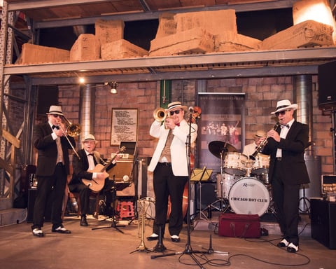 Four jazz musicians performing on stage wearing suits and fedoras, with guitar, saxophone, vocals, and drums under warm lighting